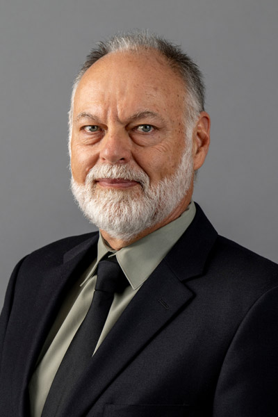 Portrait of a man with short dark hair, white beard, wearing a black blazer, black tie, and light olive green shirt.